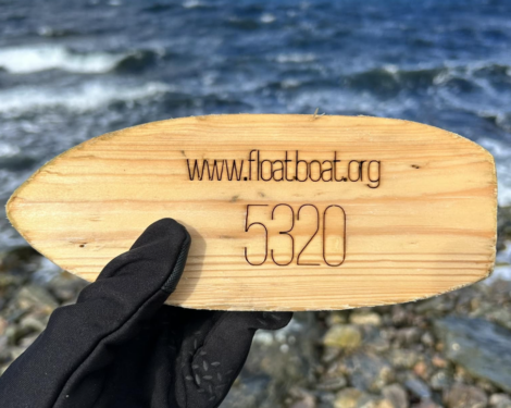 A hand wearing a glove holds an oval wooden tag engraved with "www.floatboat.org 5320" against a backdrop of a rippling body of water.