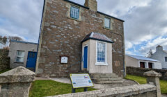 Two-story stone building with a blue door and informational sign, under a cloudy sky.