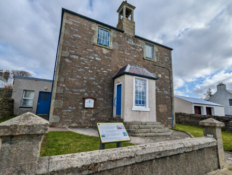 Two-story stone building with a blue door and informational sign, under a cloudy sky.