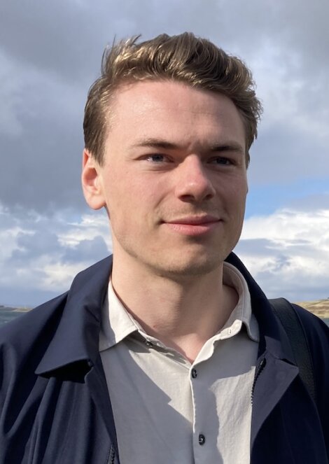 A young man with short hair standing outdoors, smiling slightly, wearing a white shirt and dark jacket against a cloudy sky backdrop.