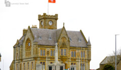 Lerwick town hall in shetland with a red flag fluttering atop, featuring a stone facade and clock tower against a cloudy sky.