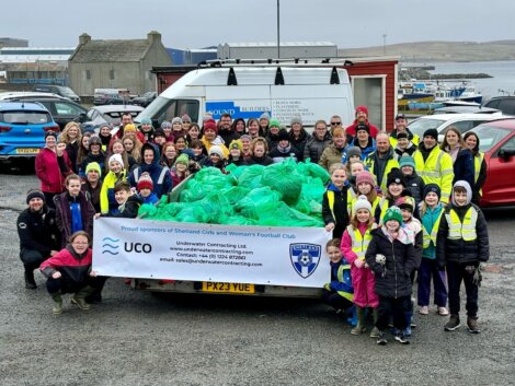 Large group of people posing with collected trash bags in front of a van with sponsorship logos, in a parking lot near water.