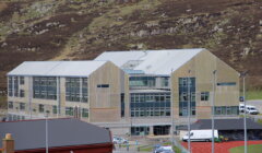 Modern wooden building with large windows located at the base of a grassy hill, featuring a parking area and adjacent red-roofed structure.