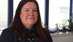A caucasian woman with shoulder-length brown hair, wearing a navy blazer and floral blouse, smiles gently, sitting in an office with blinds and plants in the background.