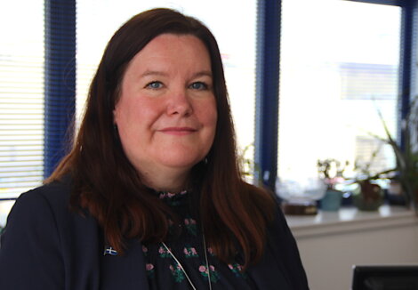 A caucasian woman with shoulder-length brown hair, wearing a navy blazer and floral blouse, smiles gently, sitting in an office with blinds and plants in the background.