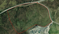 Aerial view of a rugged terrain with a red outline marking a specific area, adjacent to a road and a developed zone with buildings.