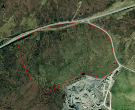 Aerial view of a rugged terrain with a red outline marking a specific area, adjacent to a road and a developed zone with buildings.