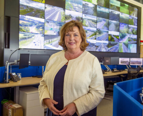 A woman stands smiling in a traffic control center, surrounded by multiple screens showing highway views.