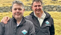 Two men in matching jackets branded with "international island games" logos, standing outdoors with a stone wall and grassy background.