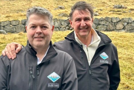 Two men in matching jackets branded with "international island games" logos, standing outdoors with a stone wall and grassy background.