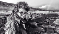 A person wearing glasses and a jacket stands beside a stone wall in a windswept landscape.