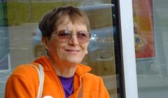 A smiling elderly woman wearing sunglasses and an orange jacket, sitting outdoors with a reflective window in the background.