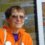 A smiling elderly woman wearing sunglasses and an orange jacket, sitting outdoors with a reflective window in the background.