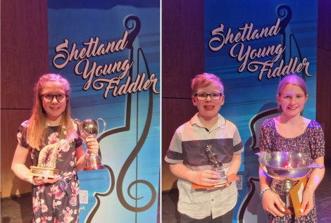 Two children holding trophies on stage at the shetland young fiddler event, with event banners behind them.