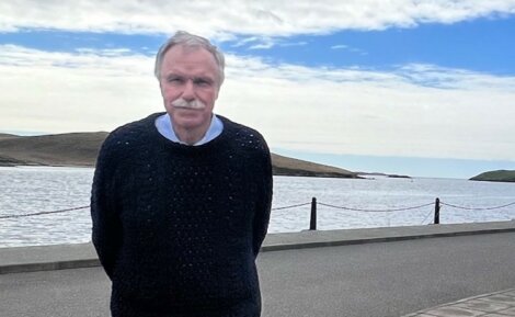 An elderly man wearing a dark sweater standing by a seaside road with a hilly island and cloudy sky in the background.