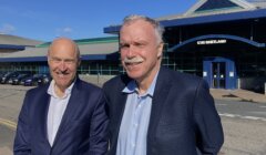 Two older men smiling in front of a building with a sign reading "uhi shetland." one is bald and wearing a dark blue suit, the other has grey hair and a grey mustache, in a light blue shirt.