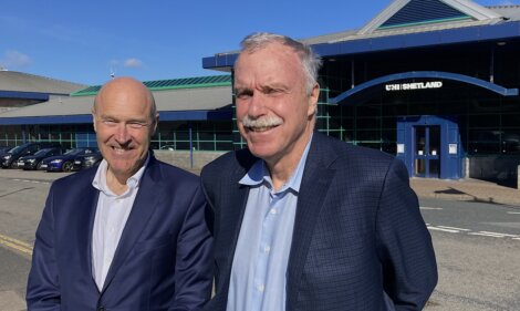 Two older men smiling in front of a building with a sign reading "uhi shetland." one is bald and wearing a dark blue suit, the other has grey hair and a grey mustache, in a light blue shirt.