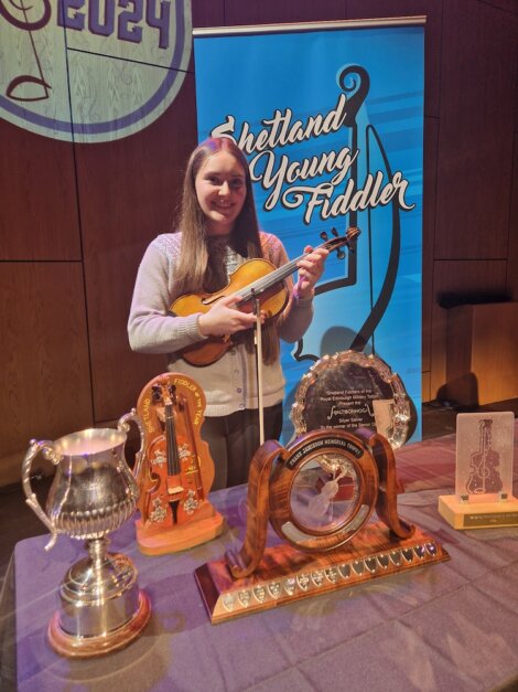 A young woman holding a violin stands beside various trophies at the shetland young fiddler event.