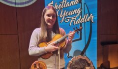 A young woman holding a violin stands in front of a banner titled "shetland young fiddler," surrounded by trophies.