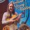 A young woman holding a violin stands in front of a banner titled "shetland young fiddler," surrounded by trophies.