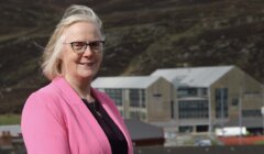 Woman in a pink blazer standing before a building with large glass windows and a mountain in the background.