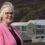 Woman in a pink blazer standing before a building with large glass windows and a mountain in the background.