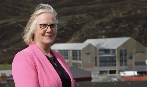 Woman in a pink blazer standing before a building with large glass windows and a mountain in the background.