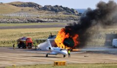 Aircraft emergency training simulation at an airport, featuring a jet with controlled fire near its engine as fire personnel oversee the exercise.