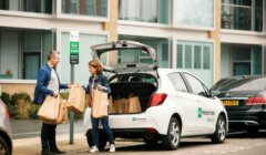 Two people exchanging shopping bags next to a car with an open trunk.