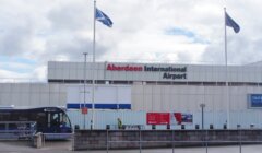 Front view of aberdeen international airport building with two scottish flags flying and a shuttle bus outside.