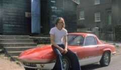 Man sitting on the hood of a red lotus elan sprint sports car, parked in front of a rustic building and garage labeled "hotz.