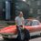 Man sitting on the hood of a red lotus elan sprint sports car, parked in front of a rustic building and garage labeled "hotz.