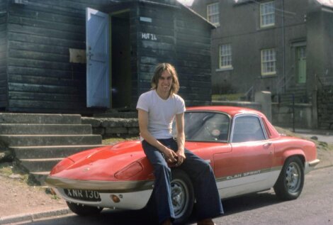 Man sitting on the hood of a red lotus elan sprint sports car, parked in front of a rustic building and garage labeled "hotz.