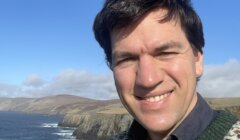 A person smiling for a selfie with a coastal landscape in the background.