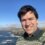 A person smiling for a selfie with a coastal landscape in the background.