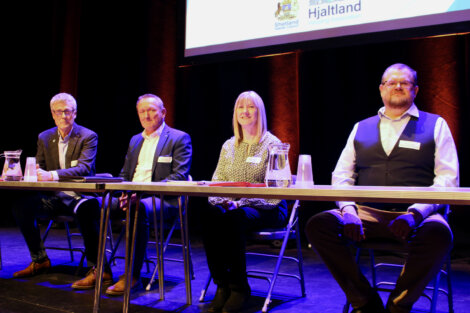 Four panelists sitting at a table on stage at a conference, with a banner reading "shetland hjaltland" in the background.