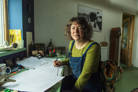 Woman sitting at a desk in a creative workspace with papers and art supplies around.