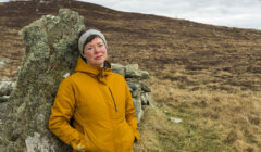 A woman in a yellow jacket resting against a rock with a grassy hill in the background.