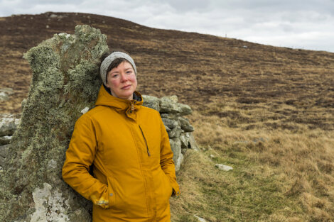 A woman in a yellow jacket resting against a rock with a grassy hill in the background.