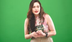 A young woman wearing a tie-dye tank top and khaki pants stands against a green backdrop, looking surprised as she checks her watch.