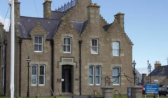 A stone civic building with a pointed roof, labeled "SOUTH RONALDSAY TOWN HALL," featuring chimneys, multiple windows, and a flagpole nearby, situated in a coastal town.