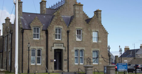 A stone civic building with a pointed roof, labeled "SOUTH RONALDSAY TOWN HALL," featuring chimneys, multiple windows, and a flagpole nearby, situated in a coastal town.