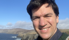 A person smiling for a selfie with a scenic coastal background on a sunny day.