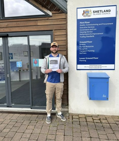 A man stands in front of a building with a sign that reads "shetland islands council," holding documents and smiling at the camera.