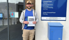 A man holding a document stands smiling in front of a building with directional signs and a blue mailbox.