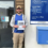 A man holding a document stands smiling in front of a building with directional signs and a blue mailbox.