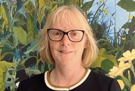 A woman with blonde hair and glasses, smiling, standing in front of a vibrant floral painting.