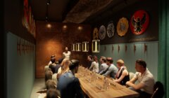 A bartender presenting to seated patrons at a dimly lit tasting room decorated with wall art and serving beer samples.