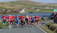 Group of runners competing in a marathon on a rural road, with houses and hills in the background.