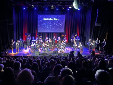 A diverse group of performers on stage at a concert, with the backdrop reading "sky full of stars" illuminated by small star-like lights, with an audience in the foreground watching.